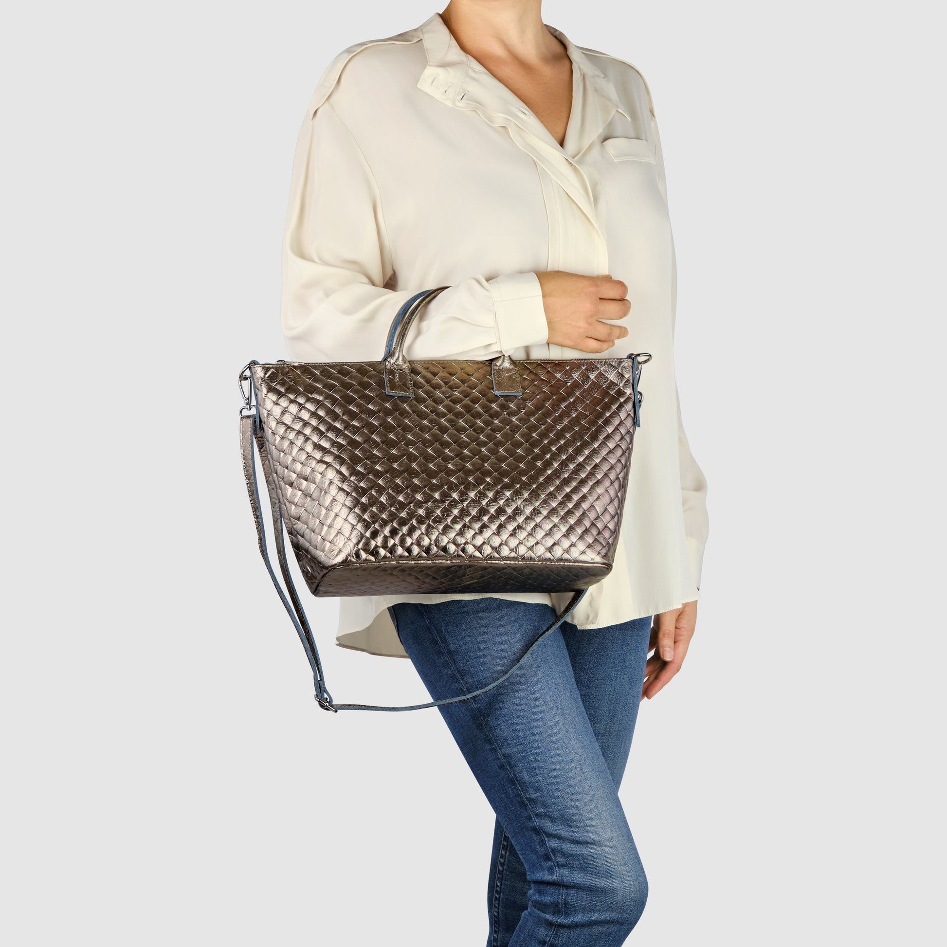 A person wearing a cream-colored blouse and blue jeans carries a large, handcrafted Italian handbag with both handles and a shoulder strap against a plain light background.