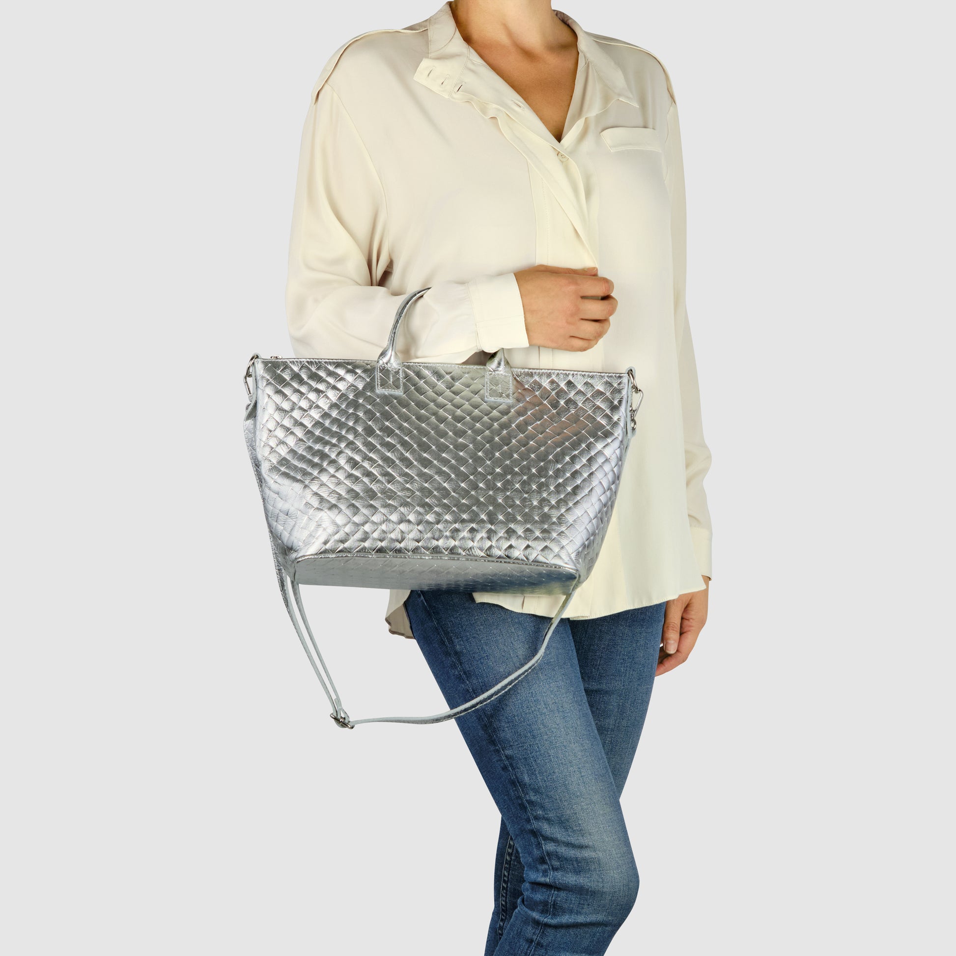 A woman wearing a cream blouse and blue jeans holds a large, handcrafted Italian handbag with both handles and a shoulder strap against a plain light background.