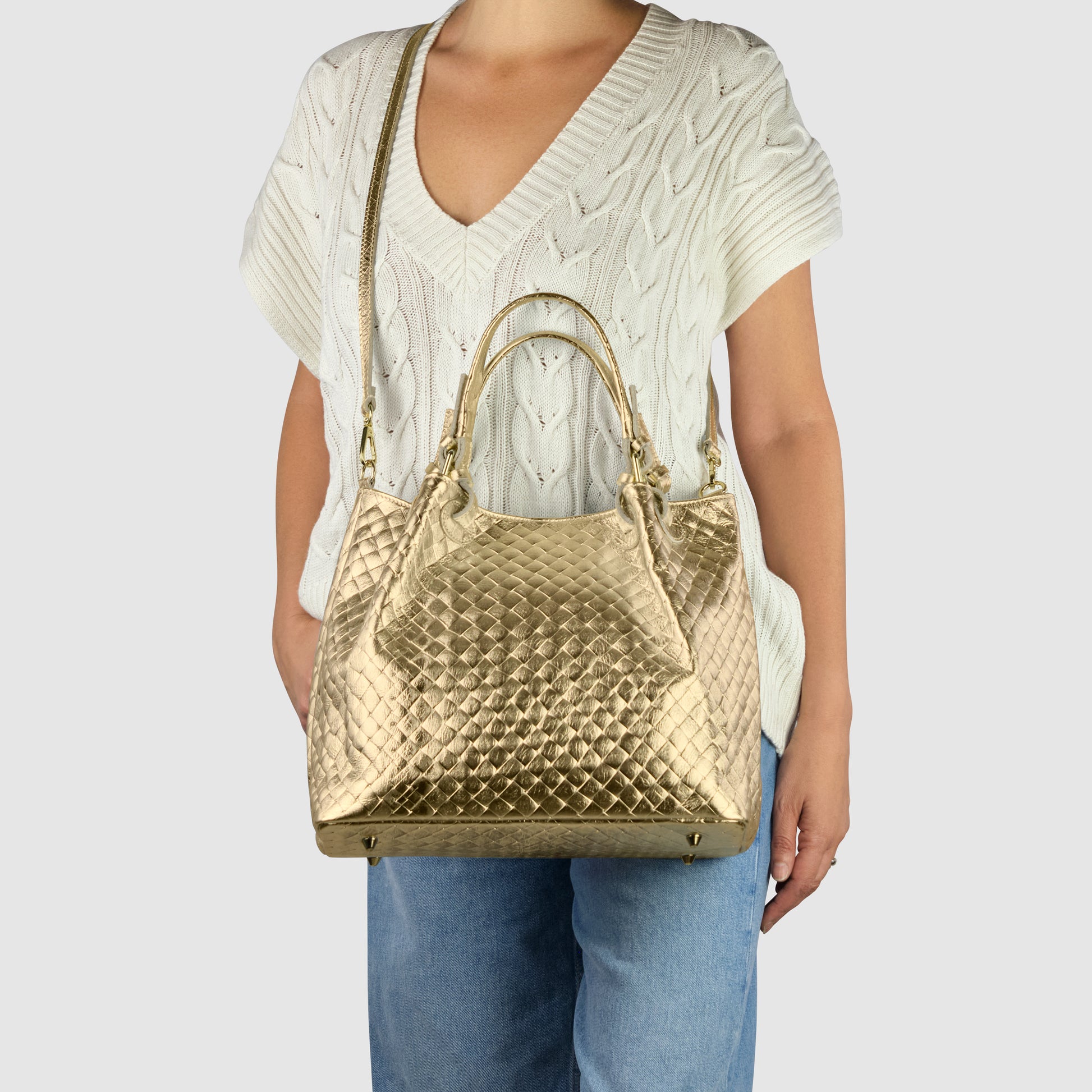 A person wearing a white short-sleeve sweater and blue jeans is holding a large, gold, Italian-made handbag with both top handles and a shoulder strap. The background is plain light gray.