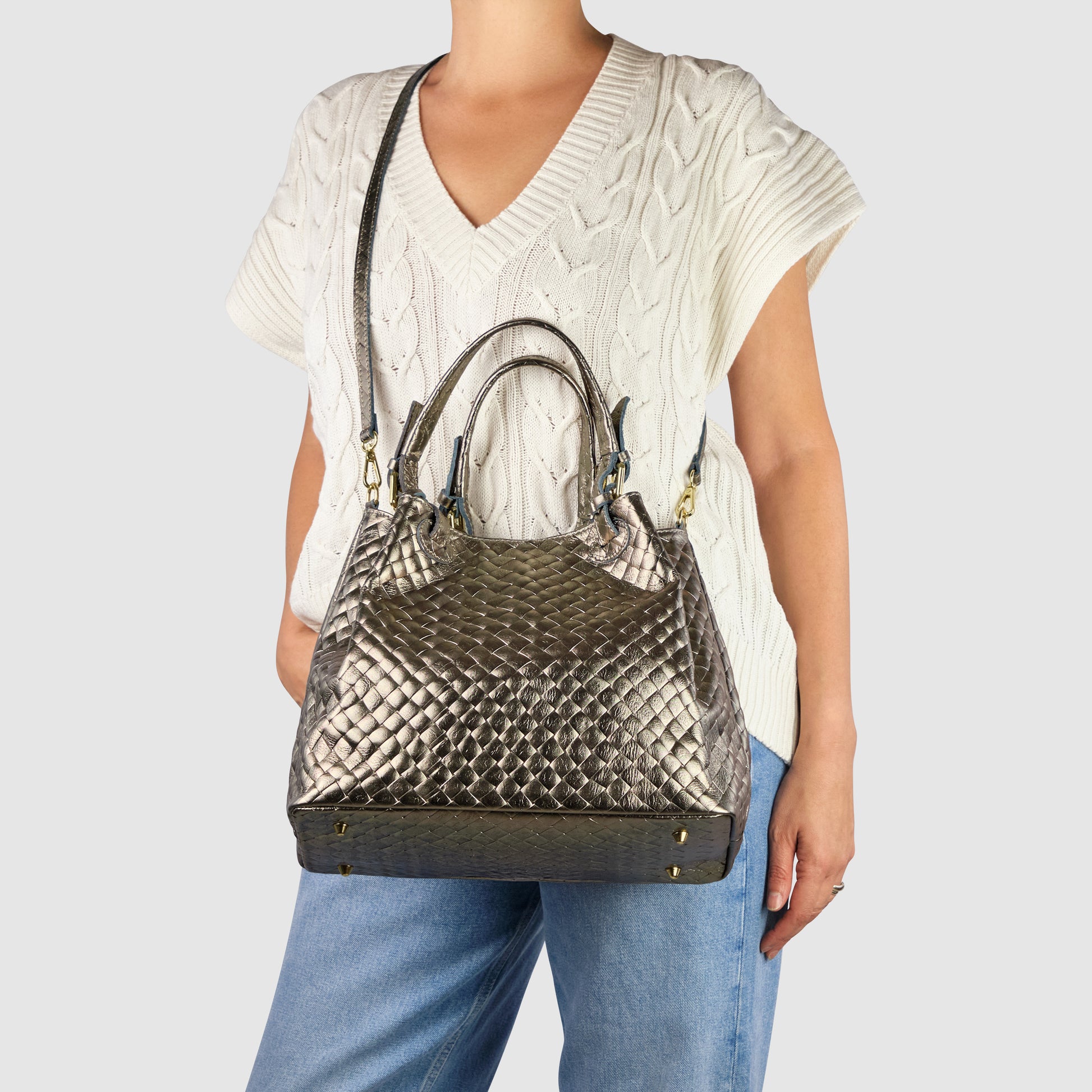 A person wearing a white cable-knit short-sleeve sweater and blue jeans holds an Italian-made metallic woven leather handbag with top handles and a shoulder strap against a plain light background.