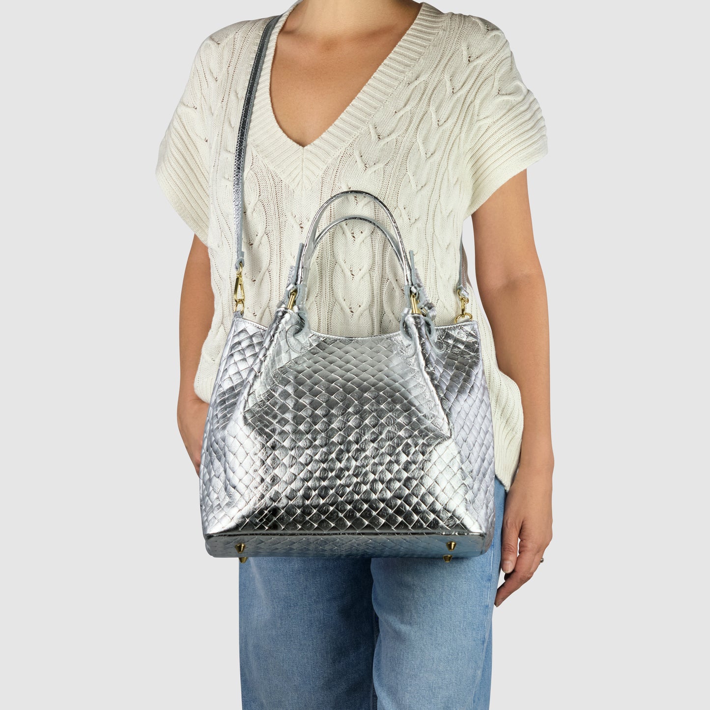 A person wearing a cream-colored, short-sleeve knit sweater and blue jeans holds an Italian-made, metallic silver quilted handbag with top handles and a shoulder strap. The bag features gold hardware and a textured pattern.