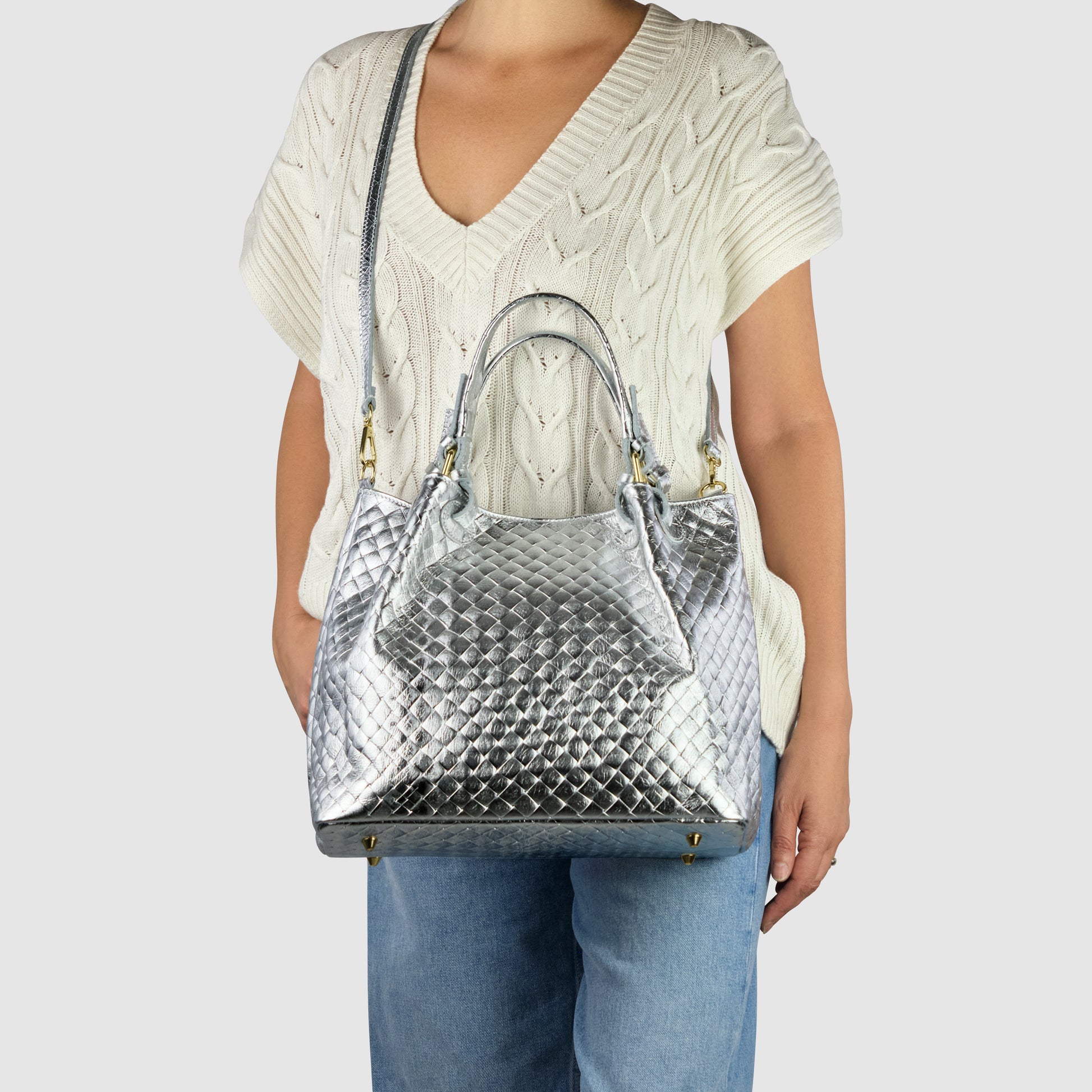 A person wearing a cream-colored, short-sleeve knit sweater and blue jeans holds an Italian-made, metallic silver quilted handbag with top handles and a shoulder strap. The bag features gold hardware and a textured pattern.