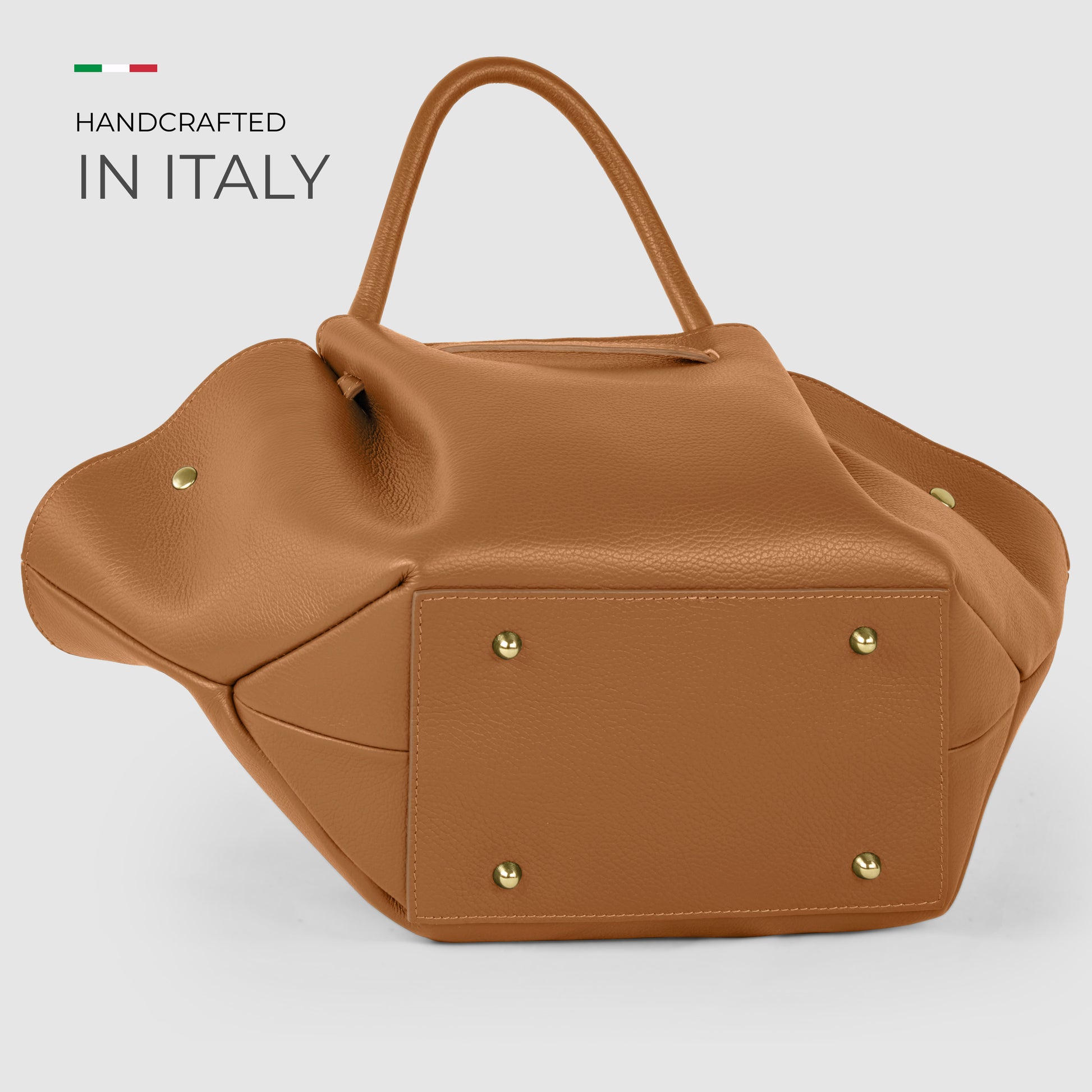 A tan Italian leather bag with gold studs on the bottom, shown against a light gray background. Text reads “Handcrafted in Italy” with a small Italian flag above the words, highlighting Florentine craftsmanship.